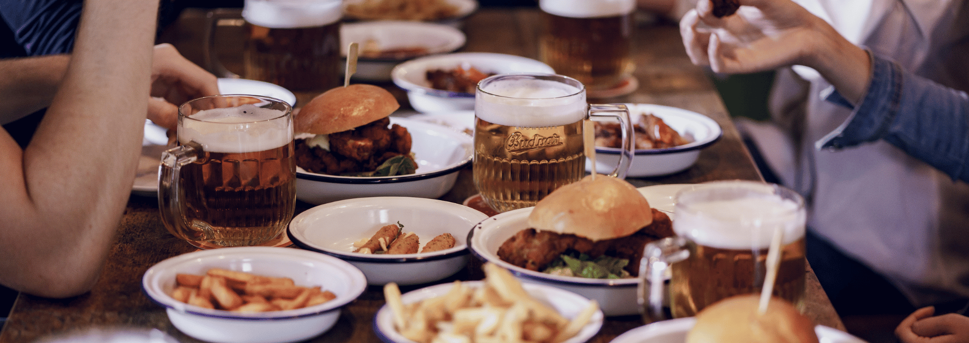 Image d'une table dans un pub où on voit des plats (burgers, frites) accompagnés de belles chopes de bières tchèques blondes provenant de la brasserie BUDWEISER BUDVAR. 
